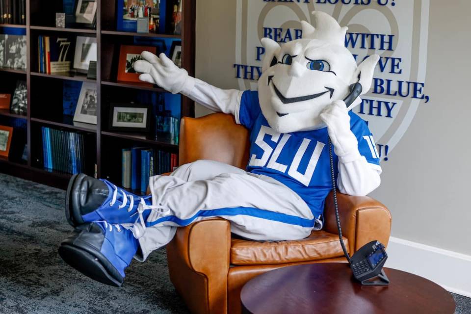 The SLU Billiken sits in a brown leather chair with his legs draped over one arm, holding a landline phone to his ear. In the background there is a bookshelf filled with many blue books and framed photographs, as well as a wall adorned with the SLU fleur de lis and the lyrics to the University's Varsity Song: "Bear we with pride and love Thy White and Blue, Sweet are thy memories, Saint ˻ֱ U!"