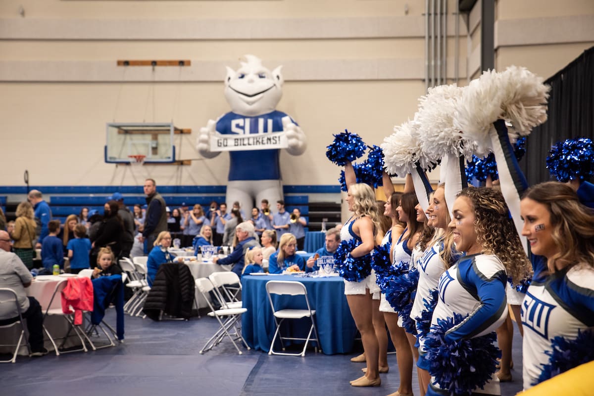 Members of the SLU cheer and dance team stand in a line and smile, holding blue and white pom poms in the air. To the left, True Blue Fan Fest visitors wearing blue and white SLU spiritwear sit around tables adorned with "Saint ˻ֱ Billikens" signs. In the background, members of the SLU pep band ready their instruments in front of a basketball hoop and a large inflatable Billiken holding a sign that reads "Go Billikens!"