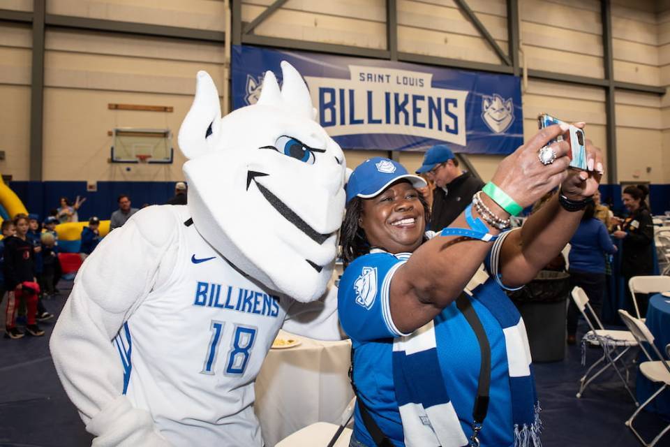 The SLU Billiken wears a white basketball jersey and poses for a selfie with a woman decked out in blue SLU spiritwear. In the background, young children play near an inflatable slide and a blue banner reading "Saint ˻ֱ Billikens" hangs on the wall. 