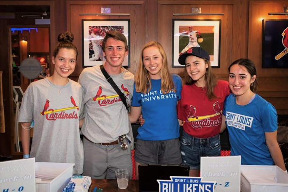 A group of five students wearing SLU and St. ˻ֱ Cardinals T-shirts pose in a wood-paneled room with framed photos of Cardinals baseball players on the wall.
